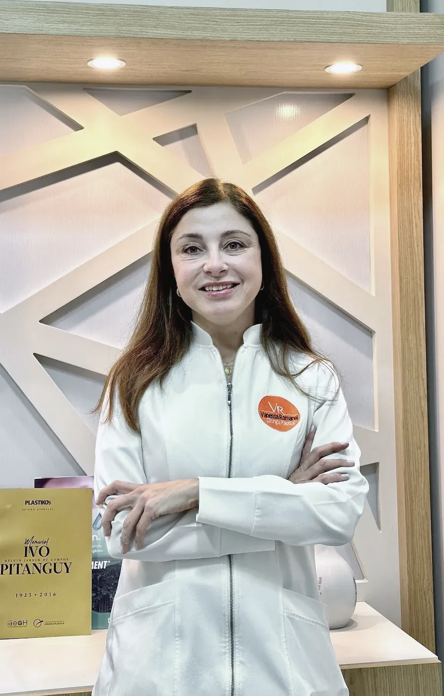 Dra. Vanessa Romanel de jaleco branco, de braços cruzados, diante da treliça iluminada do consultório